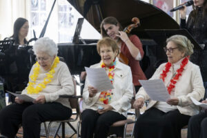 Three senior citizens sit in chairs in a row looking down at pieces of paper in their hands, as two women, a violinist and a cellist, play behind them.