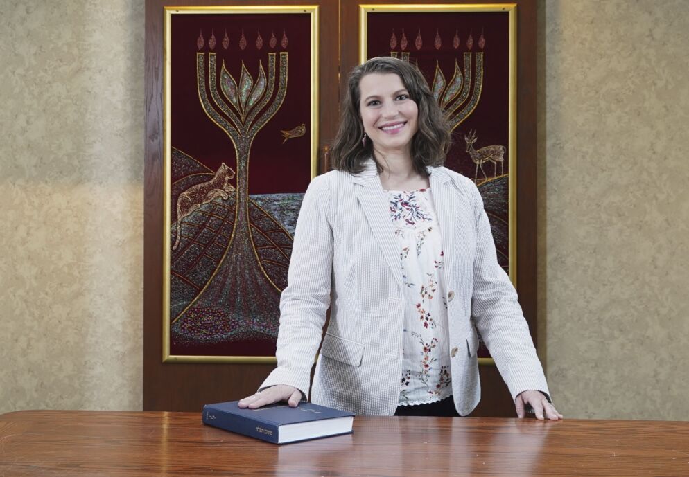Photo of a woman with shoulder-length brown hair smiling at the camera from behind a wooden desk in a sanctuary. She is wearing a cream-colored blazer over a white patterned top.