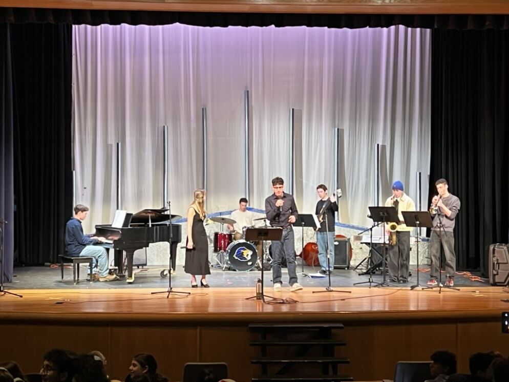 Photo of seven Israeli teens playing different instruments on stage in an ensemble. There are two vocalists, a pianist, a bass player, a drummer, a saxophone player and a trumpet player.