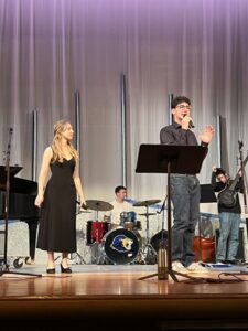 Photo of an 18-year-old boy with short dark hair and glasses singing into a microphone. A girl with long hair and a black dress looks on from next to him.