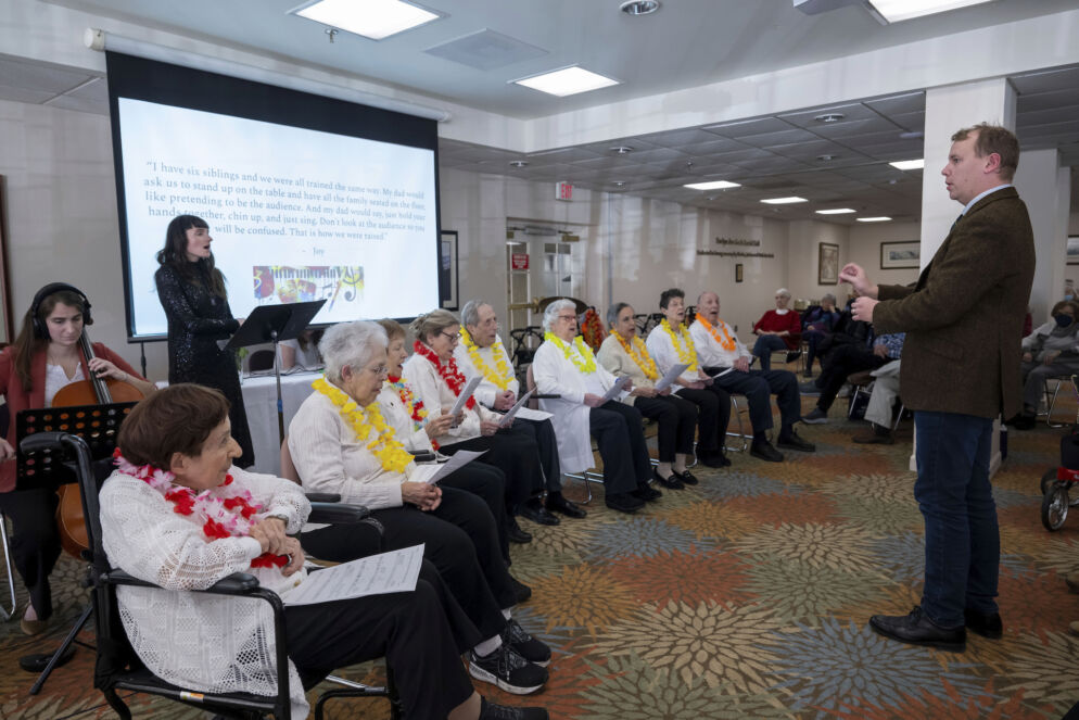 Photo of dozens of senior citizens sitting in chairs in a row in front of a man standing and facing them.