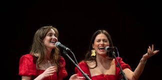 Photo of two women dressed in red singing into microphones side by side.