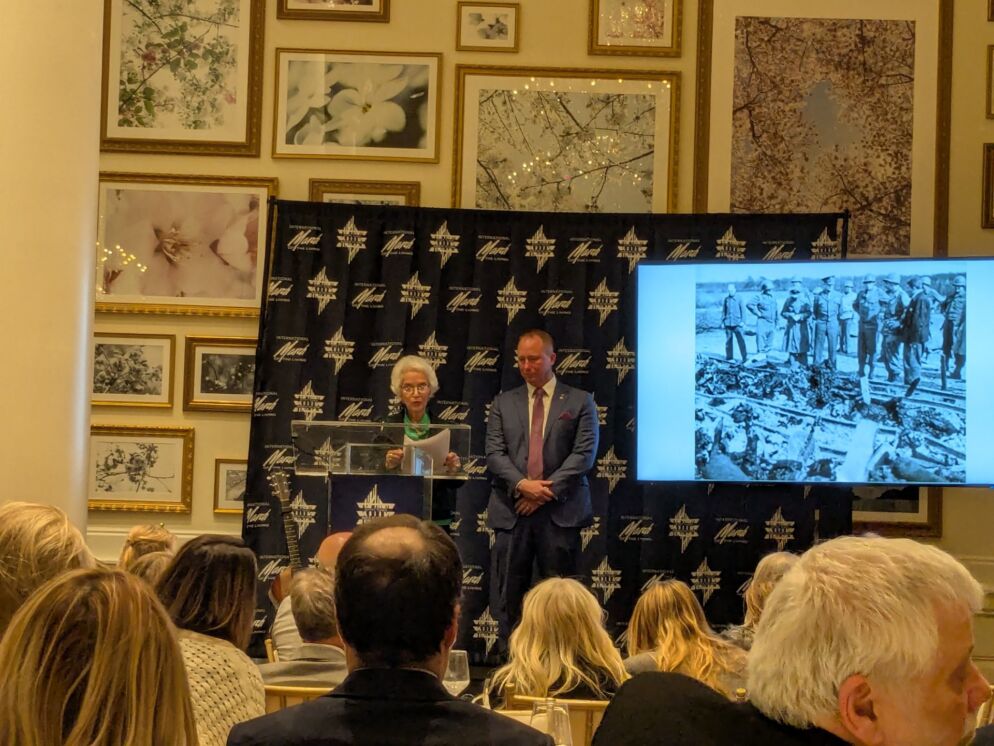 Photo of an elderly woman with short white hair and a middle-aged man in a suit standing behind a podium in front of an audience. Next to them is a screen showing a black-and-white photo of a death march from the Holocaust.