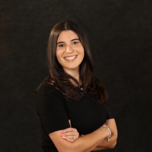 Headshot of a young woman with long straight brown hair smiling at the camera. She is wearing black.