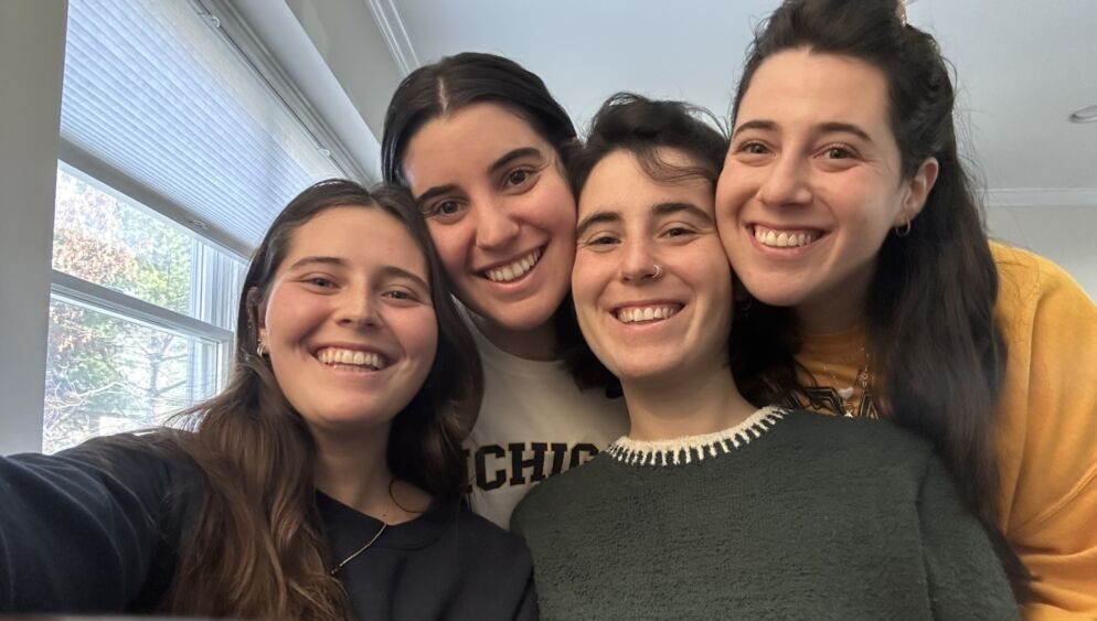 Group photo of four young women smiling at the camera from a home setting.