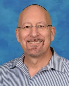 Headshot of a bald man with glasses smiling at the camera. He is wearing a black kippah and a striped button-down shirt.