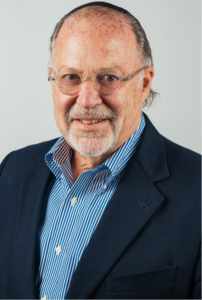 Headshot of a man with short gray hair and glasses smiling at the camera in front of a plain white background. He is wearing a black blazer  over a light blue shirt.