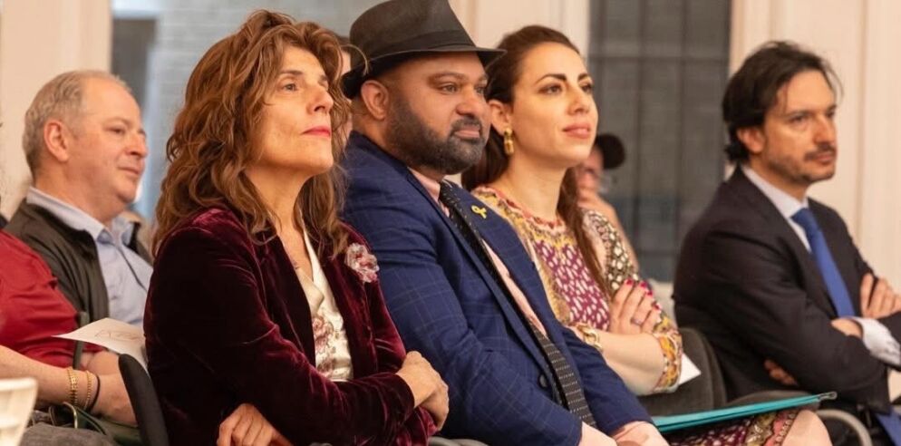 Photo of five adults seated in a concert hall. A man wearing a black fedora and a suit is seated between two women.