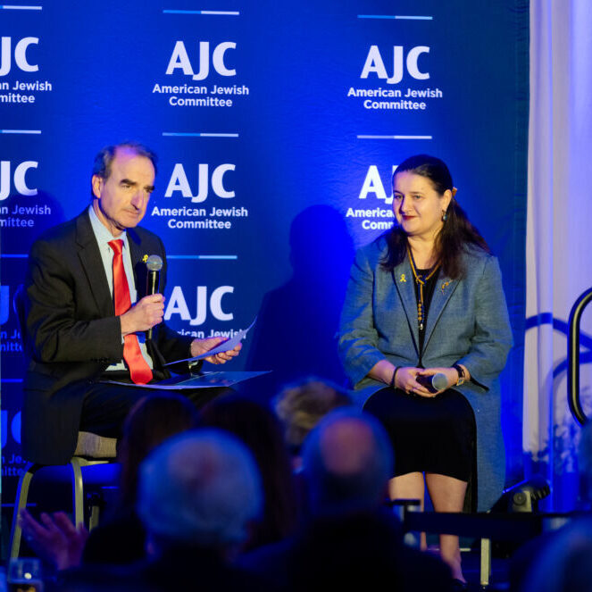 Photo of a man and a woman seated on stage with a blue banner behind them that says "AJC."