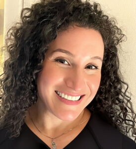 Photo of a woman with long curly dark hair smiling at the camera. She is wearing a black shirt.