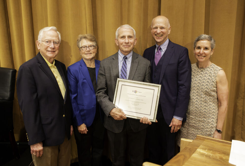 Group photo of five adults standing together dressed in suits. The man in the center is holding a framed certificate and smiling at the camera.