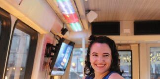 Photo of a young woman with shoulder-length dark wavy hair standing inside a Metro car refurbished as a bar. She is wearing a V-neck blue dress with a circular sticker on it and she is smiling at the camera with a hand on her hip.