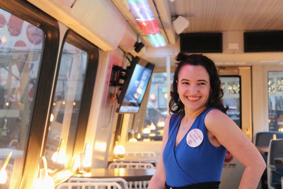 Photo of a young woman with shoulder-length dark wavy hair standing inside a Metro car refurbished as a bar. She is wearing a V-neck blue dress with a circular sticker on it and she is smiling at the camera with a hand on her hip.