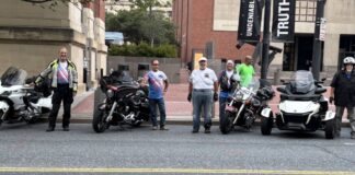 Photo of about half a dozen motorcyclists standing next to their motorcycles on the side of a city street outside a museum building.