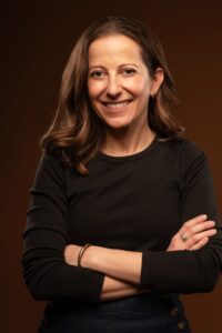 Headshot of a woman with shoulder-length brown hair. She is wearing a black long-sleeved top, crossing her arms and smiling at the camera.