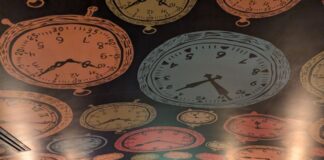 Interior of a restaurant with a colorful ceiling patterned with clock illustrations. There are red booths and small tables.