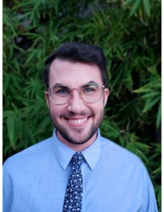 Photo of a man with short brown hair and glasses smiling at the camera. He is wearing a light blue shirt with a blue patterned tie.