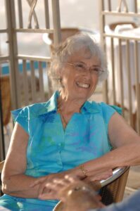 Photo of an older woman with wire-rimmed glasses and short gray hair seated in a chair. She is wearing a short-sleeved turquoise blouse and smiling.