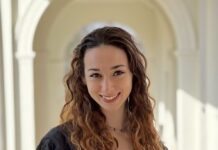 Headshot of a young woman with long wavy brown hair. She is wearing a black robe over a white top.