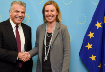 Yapir Lapid and Federica Mogherini shake hands next to a European Union flag.
