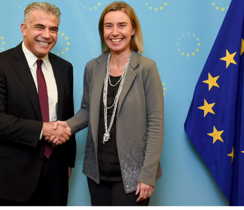 Yapir Lapid and Federica Mogherini shake hands next to a European Union flag.