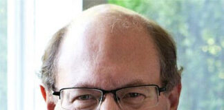 Photo of a balding man with glasses sitting in front of a living room window. He is smiling at the camera.