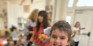Photo of a young girl wearing pajamas in a classroom and holding a pastel rainbow-colored stuffed animal. She has very short brown hair and is smiling at the camera.
