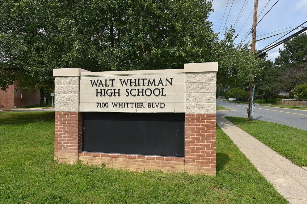 Photo of a brick entrance sign that reads "Walt Whitman High School" in black serif type. Below that, in smaller text reads "7100 Whittier Blvd." There is a sidewalk, trees and grass in the background.