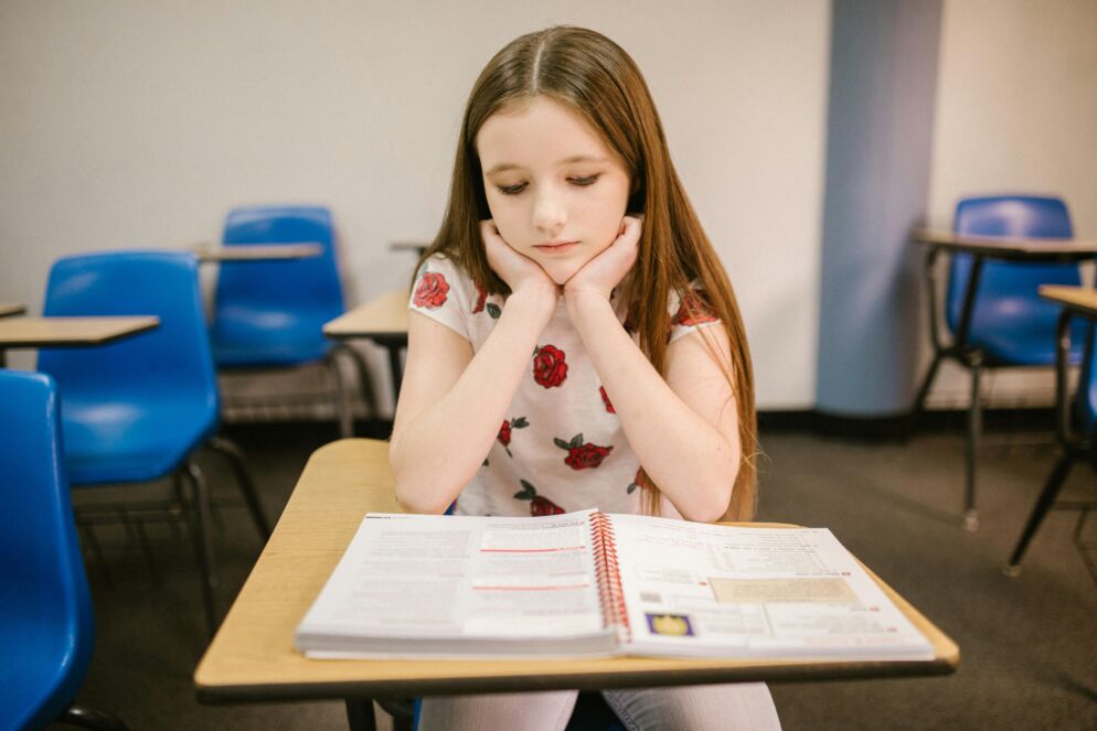 Stock photo of a young adolescent girl with long straight brown hair sitting by herself at a classroom desk. She is resting her chin in her hands with her elbows on the desk reading a book solemnly. She is wearing a white T-shirt with a pattern of red roses.