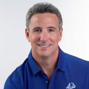 Headshot of a middle-aged man with short gray hair smiling at the camera against a white background. He is wearing a blue collared shirt.