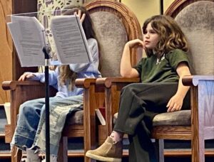 Photo of two kids sitting in upholstered chairs in a synagogue sanctuary looking at a music stand containing a paper script.