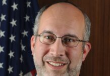 Headshot of a man with short gray hair wearing a suit and tie and glasses. He is smiling at the camera.
