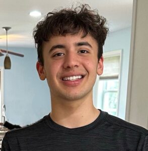 Selfie of a high school-age boy with short wavy brown hair. He is wearing a dark gray shirt and smiling at the camera.