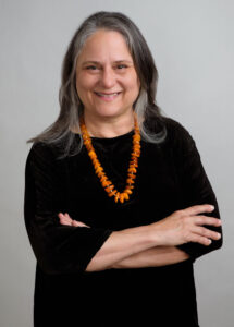 Headshot of a woman with shoulder-length straight gray hair. She is smiling at the camera with her arms crossed in front of her. She is wearing a black top with a large beaded orange necklace.