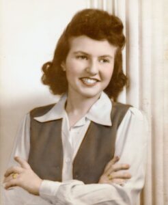 Sepia-toned photo of a young woman with chin-length wavy brown hair smiling with her arms crossed in front of her. She is wearing a gray vest over a collared white shirt.